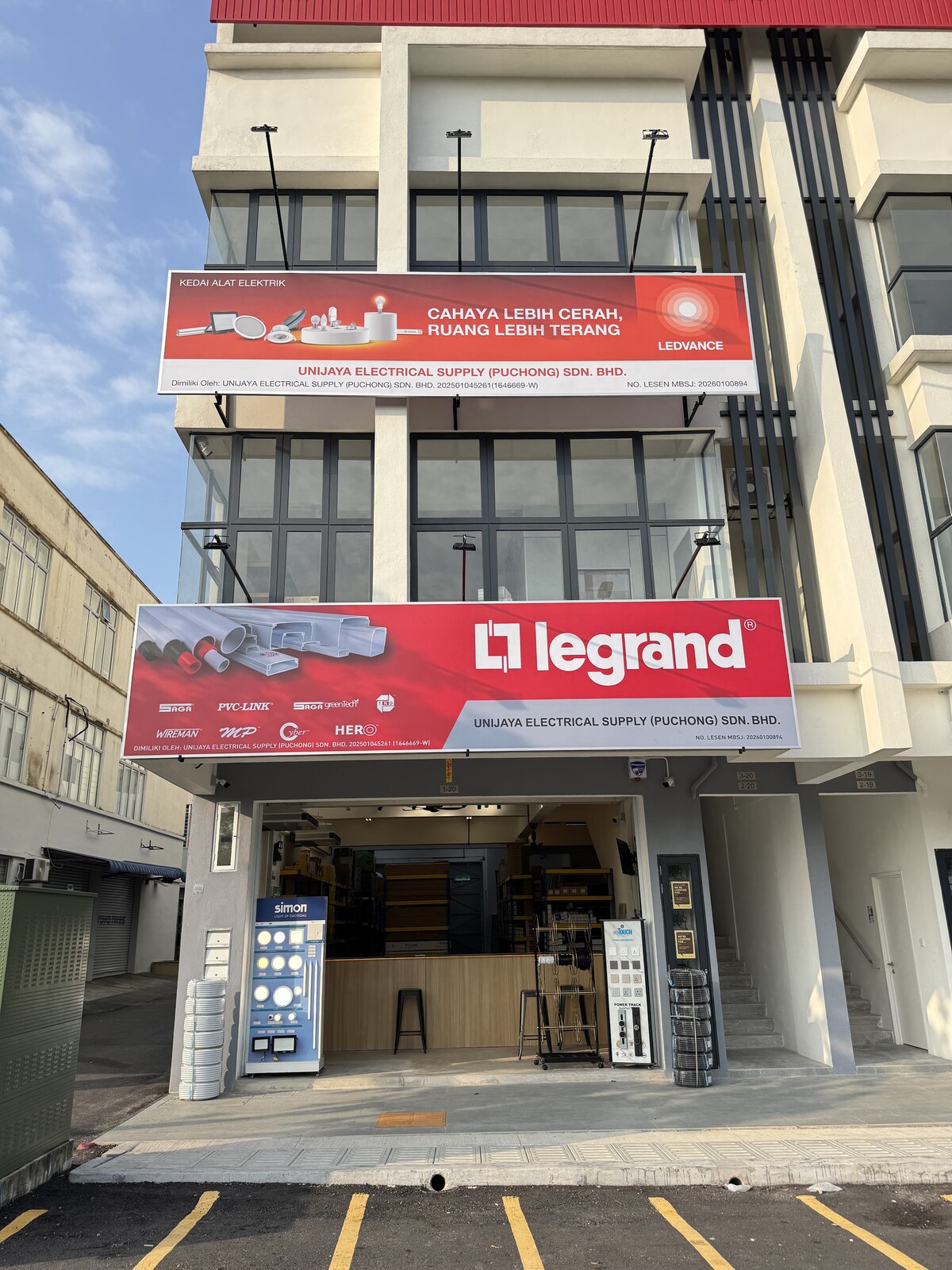 Four-storey corner block with a red Ledvance signboard and a red Legrand signboard above an open shopfront with a counter, display stands and coils of conduit