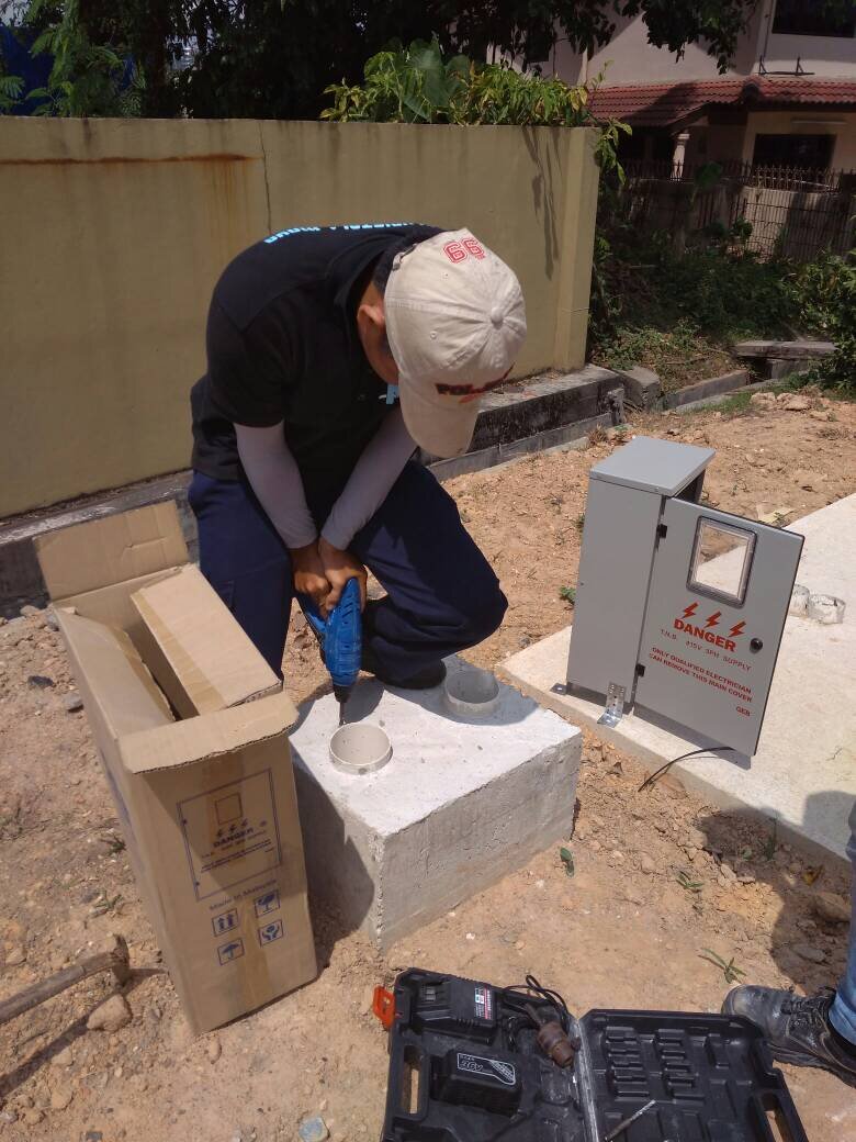 Technician in a cap drilling into a concrete plinth beside a grey outdoor meter panel marked Danger 415V 3PH supply, with the panel's carton and a tool case on the ground