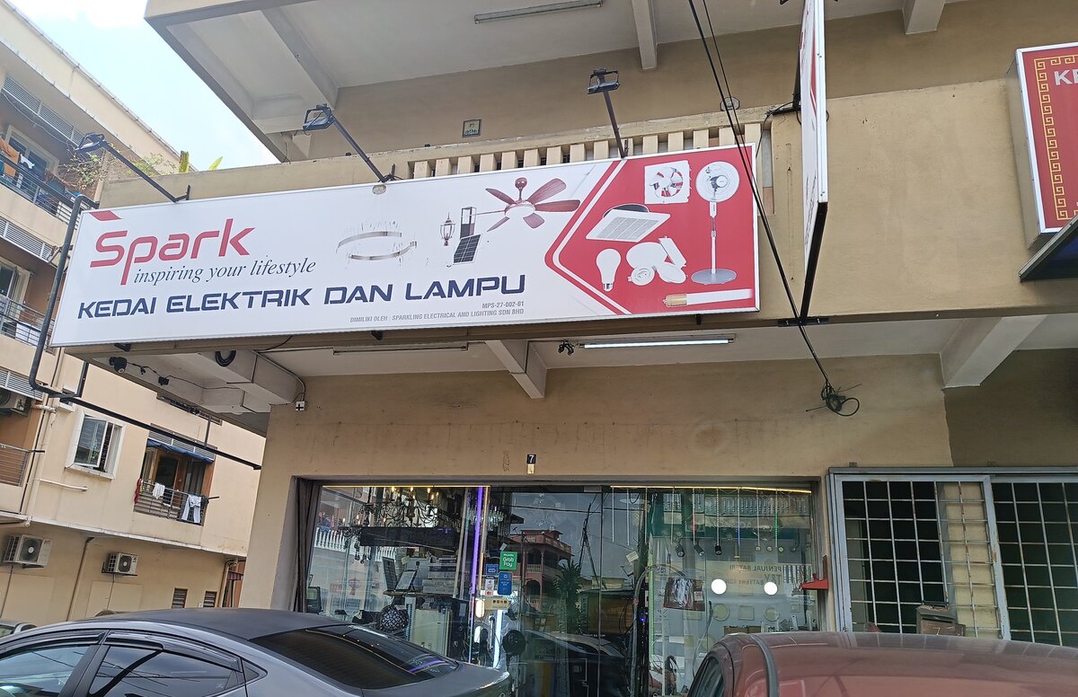 White and red Spark signboard reading Kedai Elektrik dan Lampu above a glass shop front with chandeliers and lights