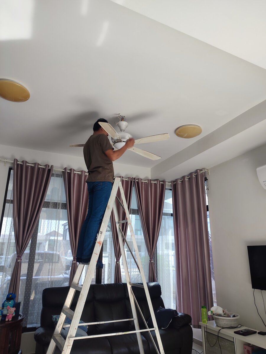 Technician on an aluminium ladder installing a wooden-blade ceiling fan in a living room with curtains and a sofa