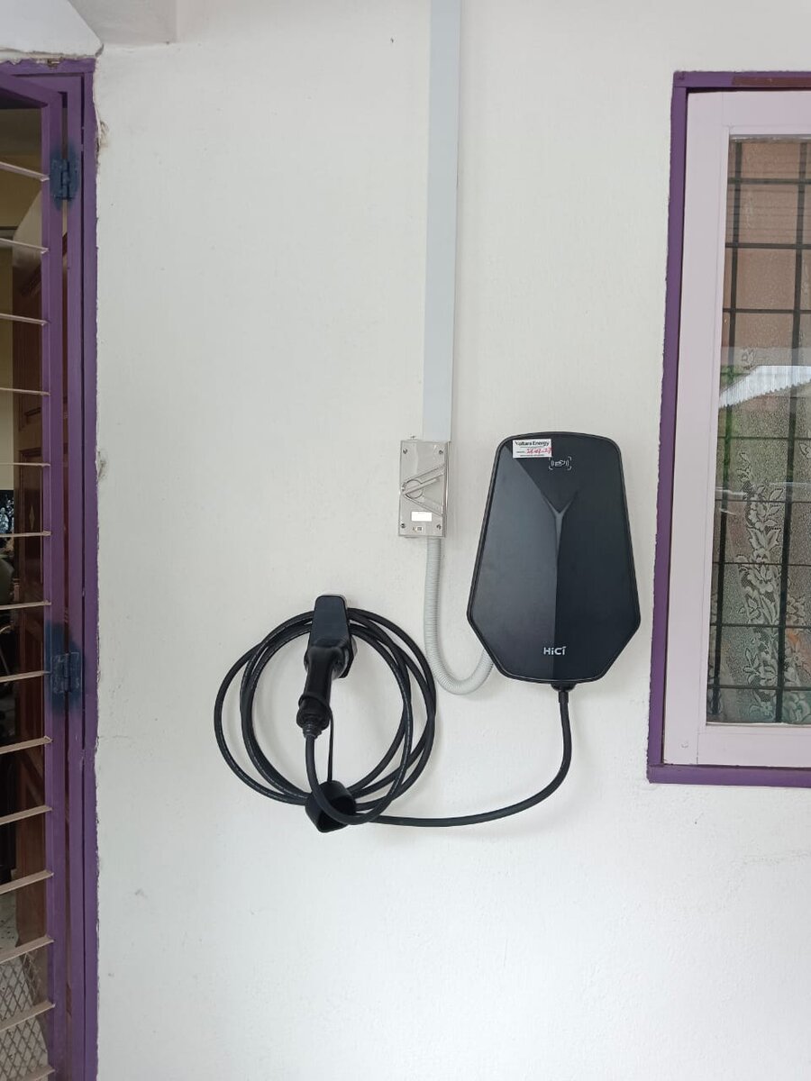 Black wall-mounted electric-vehicle charger with a coiled charging cable on a white porch wall, fed from a switch through flexible conduit and white trunking