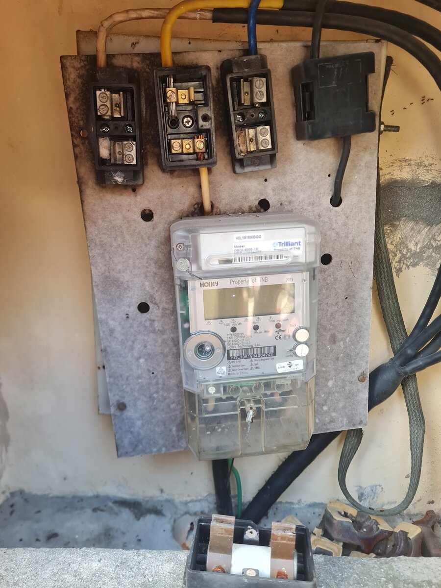 Weathered meter board with three black cut-out fuse holders and a neutral link above a digital smart meter, with thick black cables and a removed fuse carrier at the bottom