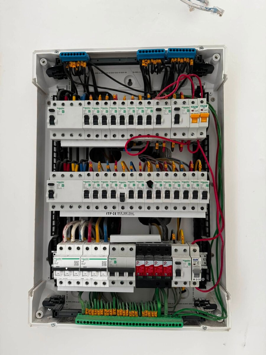 Open surface-mounted distribution board with three rows of white circuit breakers, red surge-protector modules, neatly dressed coloured wires with yellow labels and a green earth bar