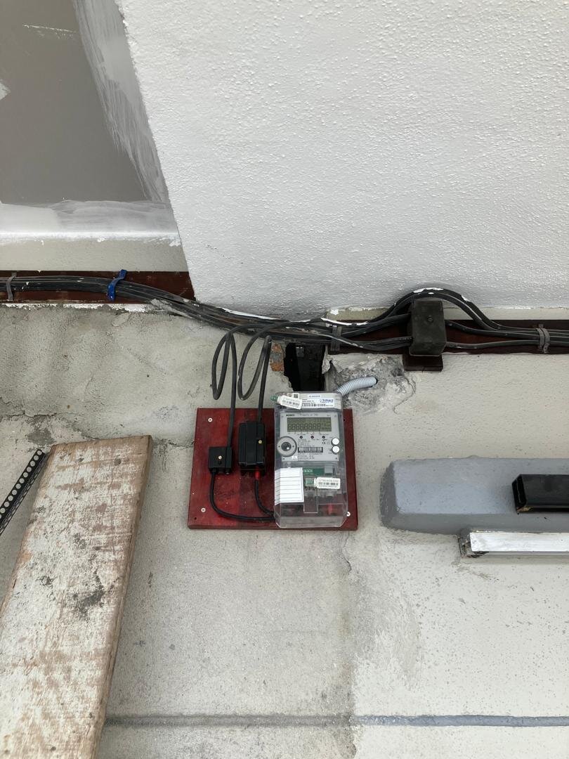 Digital single-phase electricity meter and two black cut-out fuse carriers mounted on a red board under a concrete overhang, with cables run along the wall above