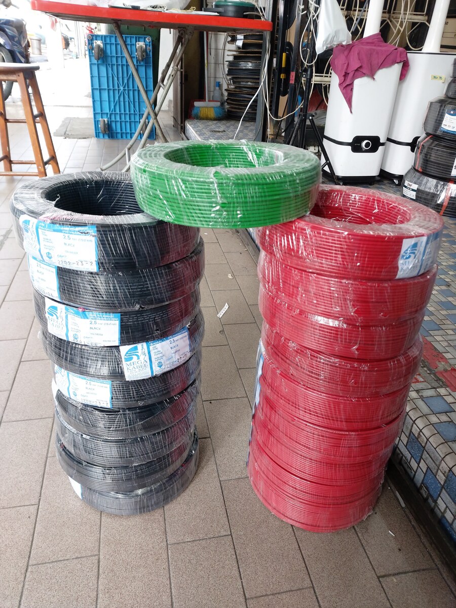 Two stacks of plastic-wrapped black and red cable coils with Mega Kabel labels and a green coil on top, on a tiled walkway in front of a shop