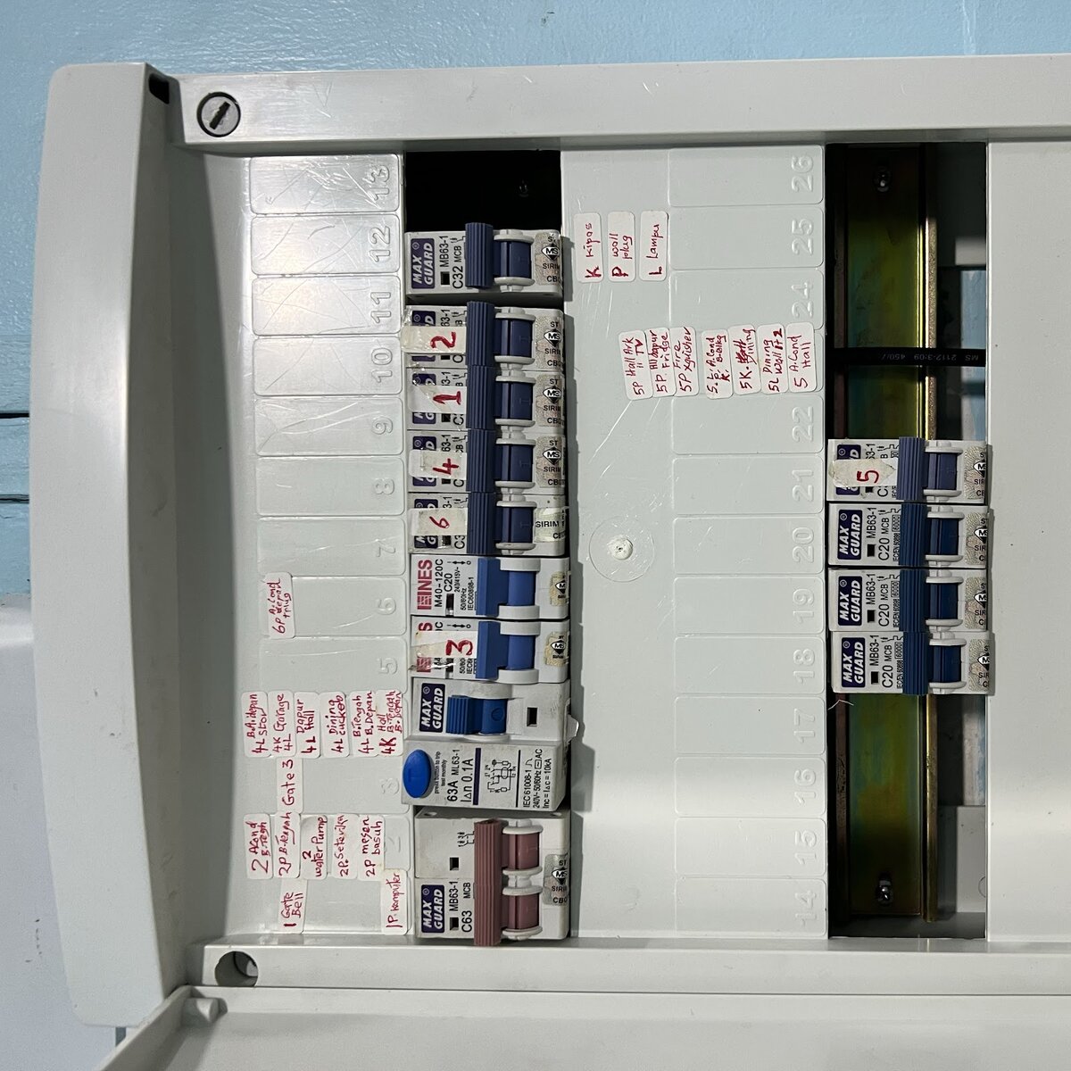 Open grey distribution board with two columns of blue miniature circuit breakers, an RCCB with a blue test button and small handwritten labels for fans, sockets, lights, aircond and water pump