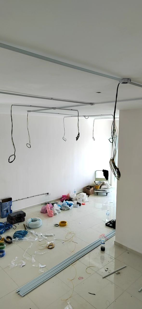 Empty room with white plastic conduit fixed across the ceiling, loops of cable hanging from junction points, and coils of cable and conduit lengths on the tiled floor