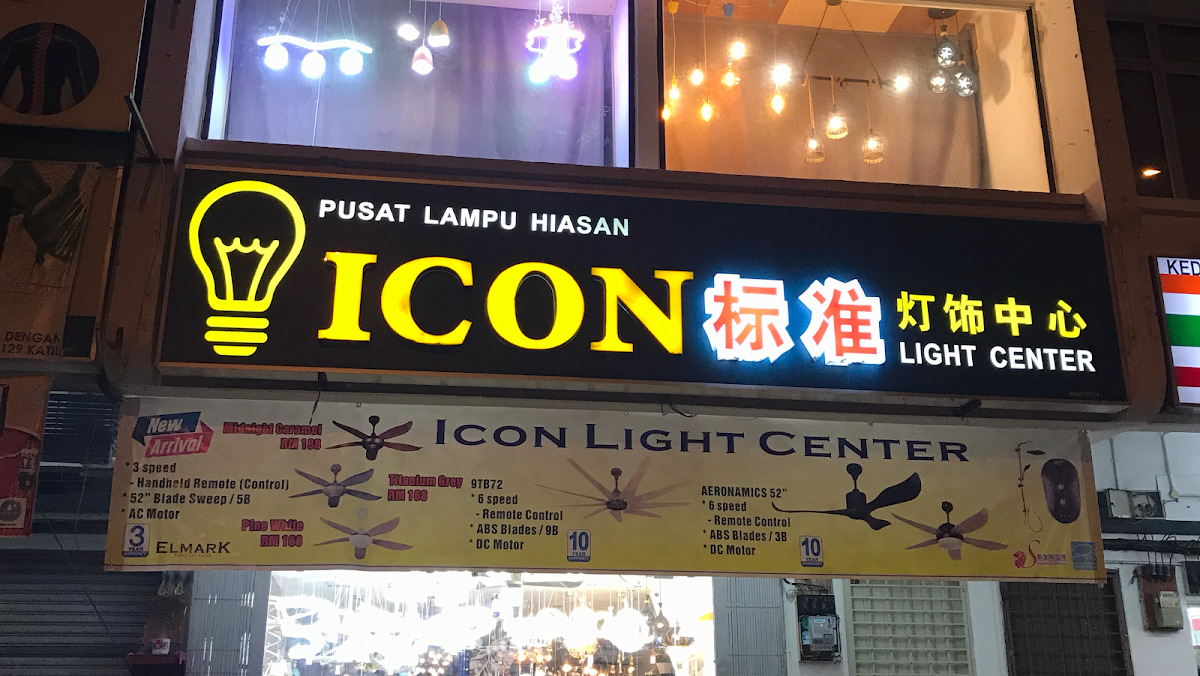Illuminated black shop sign with a yellow light-bulb logo reading Pusat Lampu Hiasan, Icon, Light Center, above a yellow banner advertising ceiling fans with prices
