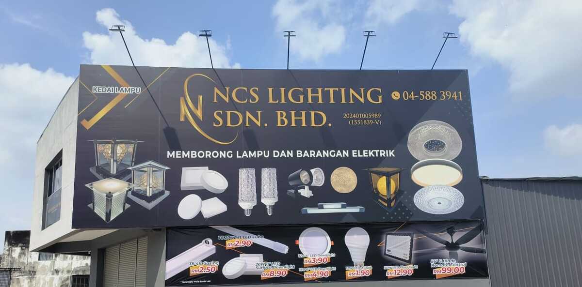 Large black and gold billboard on a corner building reading NCS Lighting Sdn Bhd with phone number and pictures of gate lamps and LED lights, above a banner of lighting products with red price tags