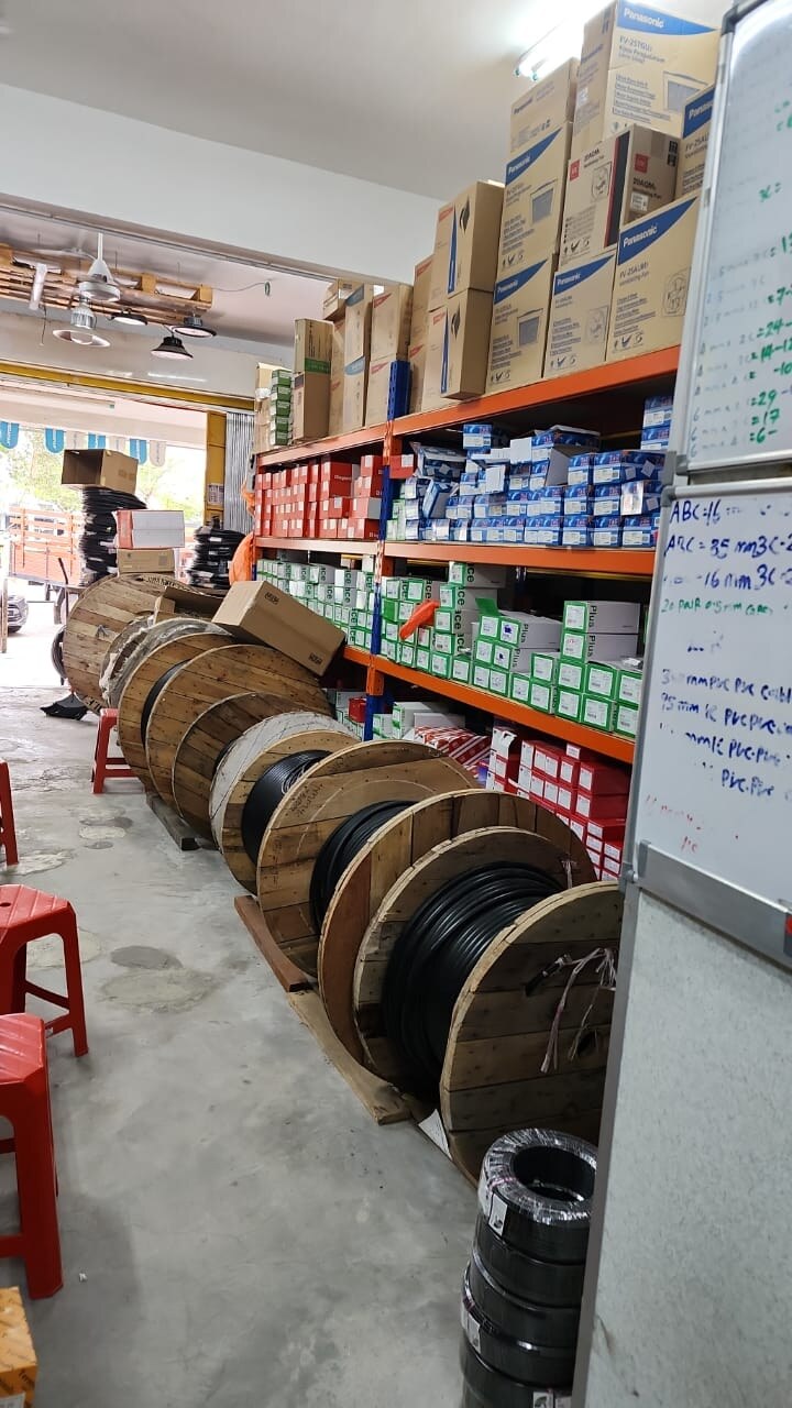 Narrow shop aisle with wooden cable drums of black cable along the floor, orange shelving stacked with boxed electrical goods, and a whiteboard of handwritten cable orders