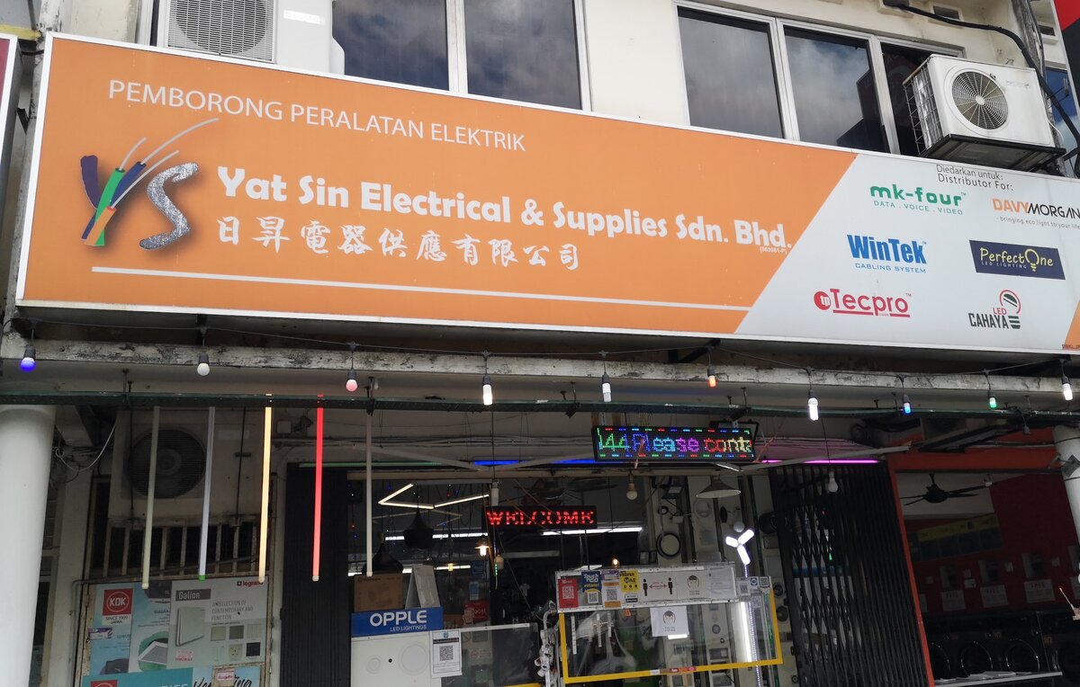 Orange Yat Sin Electrical & Supplies signboard with Chinese characters and a white panel of brand logos above an open shopfront hung with lamps, with KDK and Legrand posters on the left