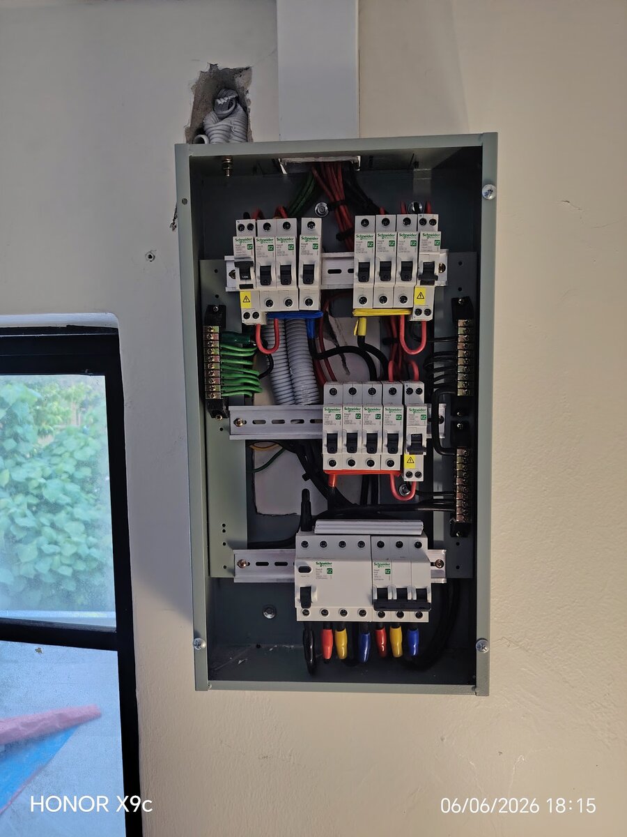 Open grey distribution board on a wall with three rows of white Schneider miniature circuit breakers and residual-current breakers, earth and neutral bars at the sides and a four-pole RCCB and three-pole main breaker on the bottom rail