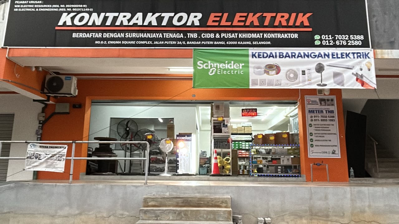 Black signboard reading Kontraktor Elektrik with WhatsApp numbers above an orange shopfront, a green Schneider Electric banner reading Kedai Barangan Elektrik, fans and shelves of electrical goods inside
