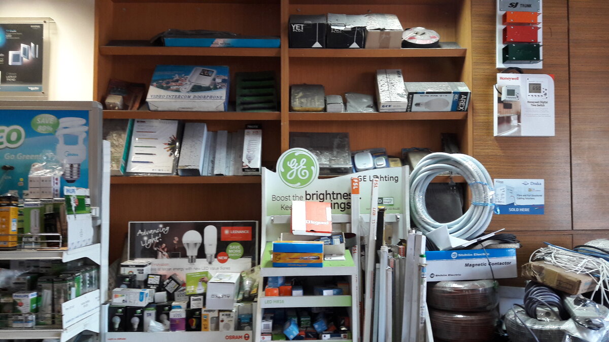 Wooden shelves of boxed lamps and electrical parts behind a GE Lighting display stand, a Ledvance lamp display, a coil of white flexible conduit and wrapped cable coils