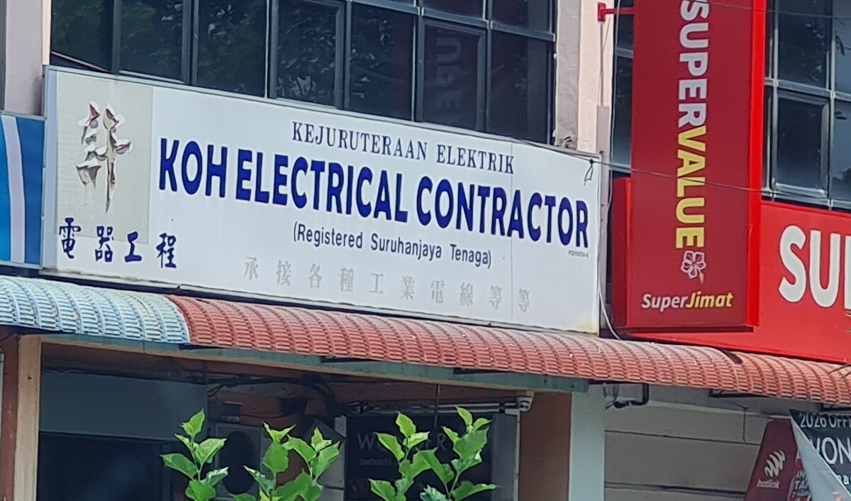 White shopfront signboard reading Kejuruteraan Elektrik, Koh Electrical Contractor, Registered Suruhanjaya Tenaga, with Chinese characters, above a shop with a red-and-white awning; a supermarket sign is next door