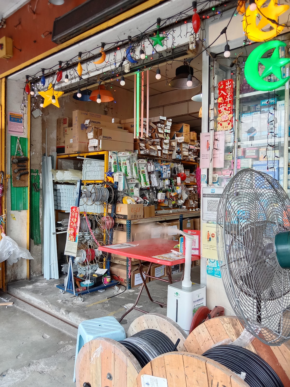 Open shopfront with cable drums on the floor, a stand fan and shelves of electrical fittings