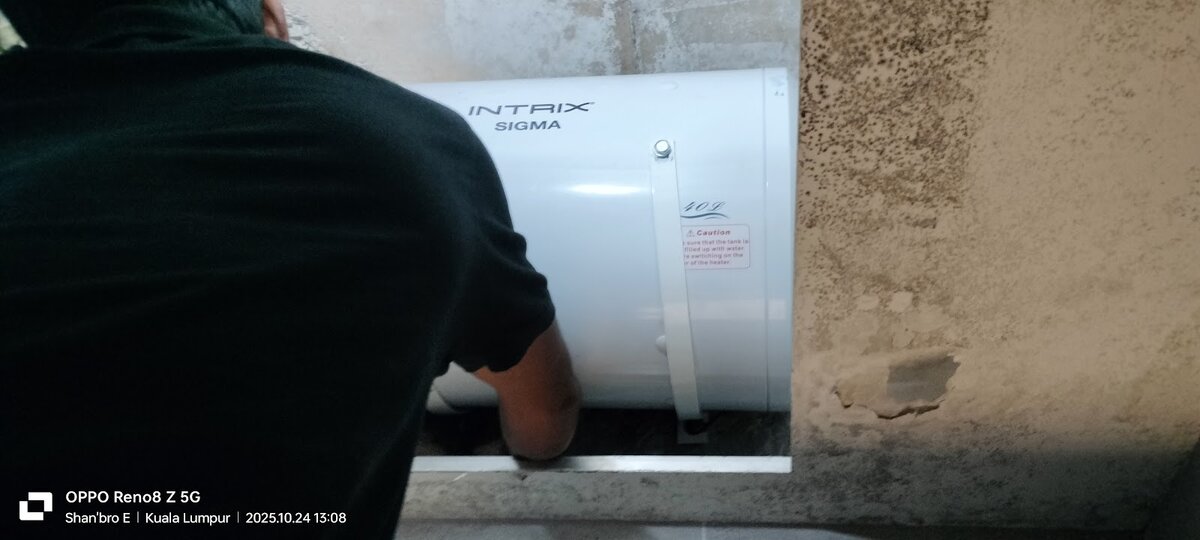 A man in a black T-shirt reaching behind a white Intrix Sigma storage water heater mounted near a ceiling, with a stained wall beside it