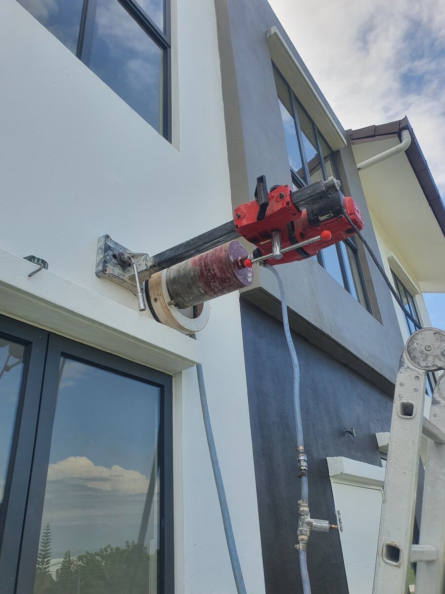 Red core-drilling motor on a steel column bolted to a white exterior wall above a window, with a water hose attached