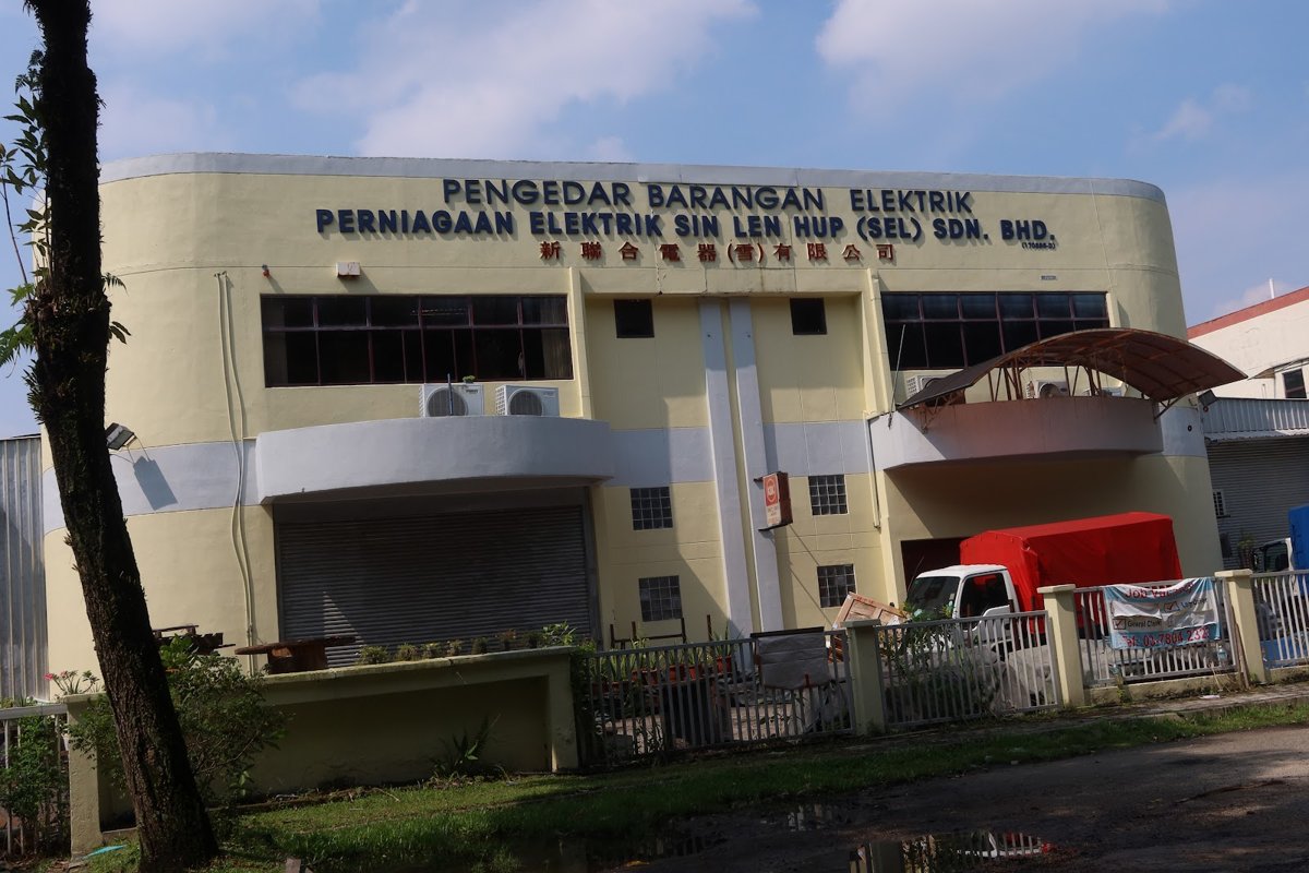 Two-storey cream building with blue lettering reading Pengedar Barangan Elektrik and Perniagaan Elektrik Sin Len Hup (Sel) Sdn. Bhd. with Chinese characters, a roller shutter and a red-covered lorry behind a white fence