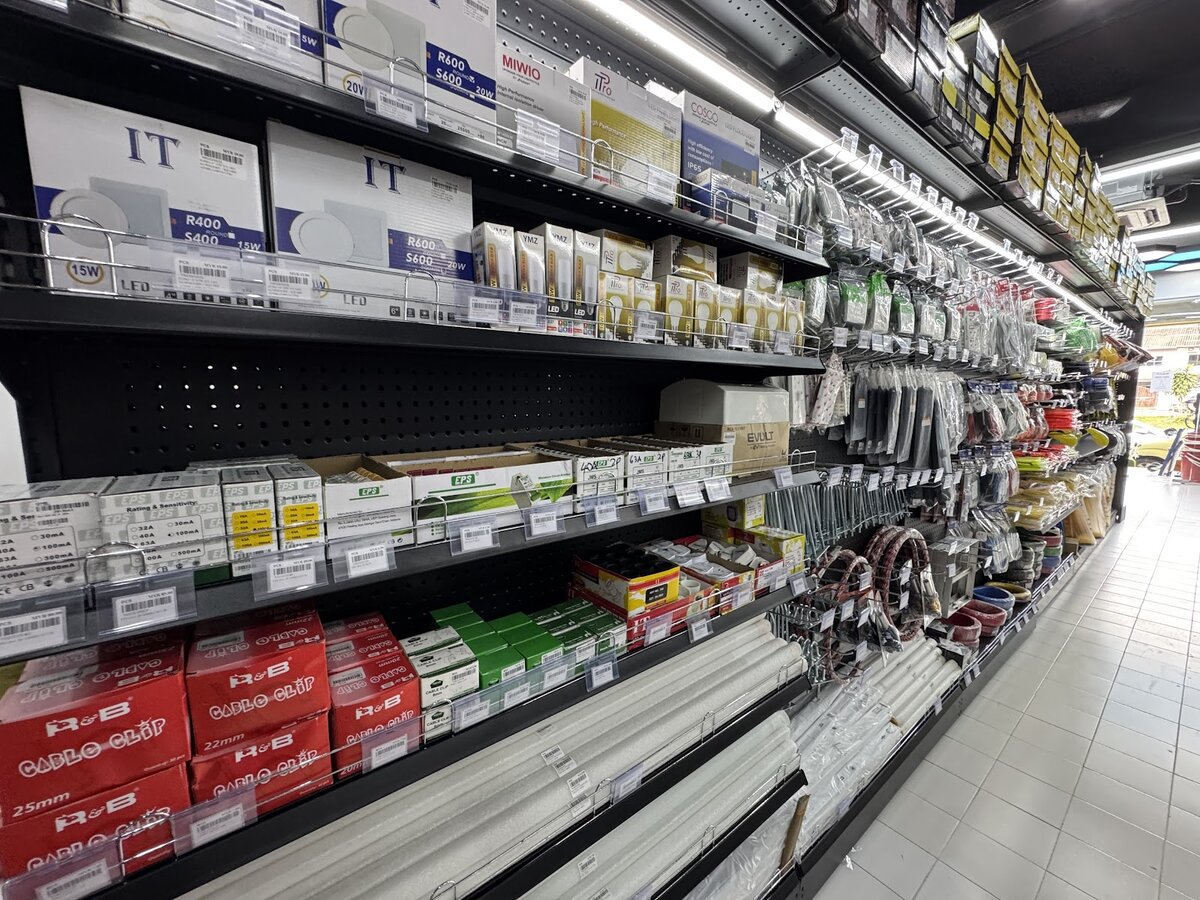 Long shop aisle with boxed LED downlights and bulbs on the top shelves, circuit breaker boxes and cable-clip cartons below, PVC conduit lengths and coils of cable along the bottom