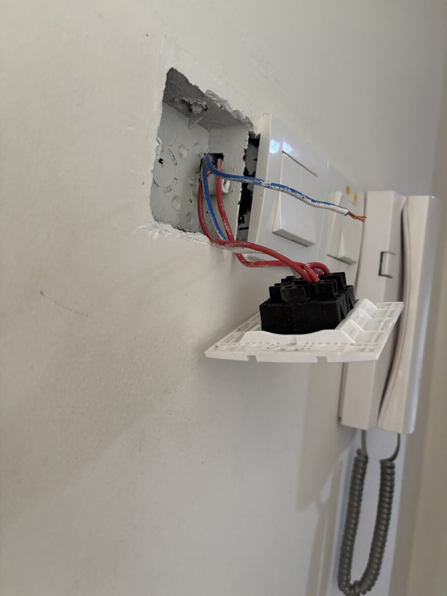 Wall switch plate pulled out of its box showing red and blue wires, next to a white intercom handset on a white wall