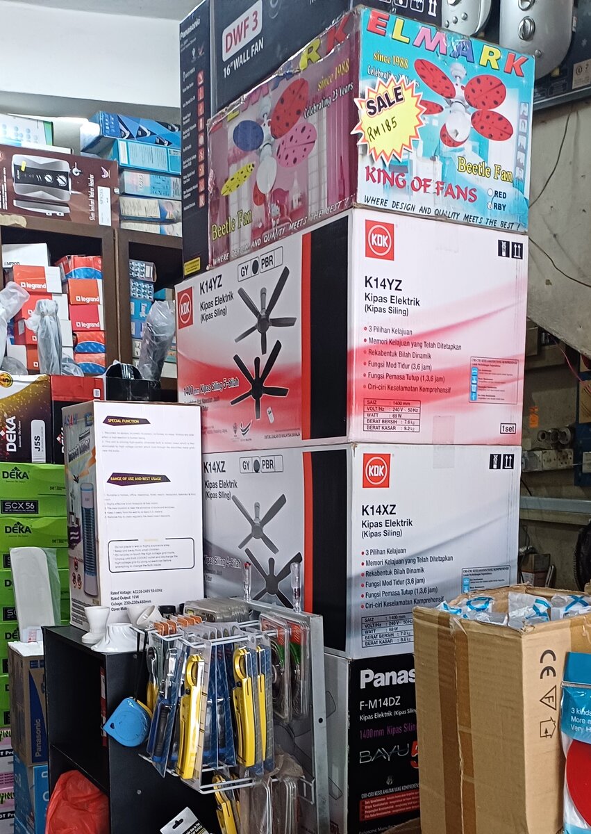 Stacked ceiling-fan cartons labelled KDK K14YZ, KDK K14XZ and Panasonic F-M14DZ beside an Elmark fan box, with green Deka boxes and red Legrand boxes on shelves and a hanging rack of yellow cable cutters
