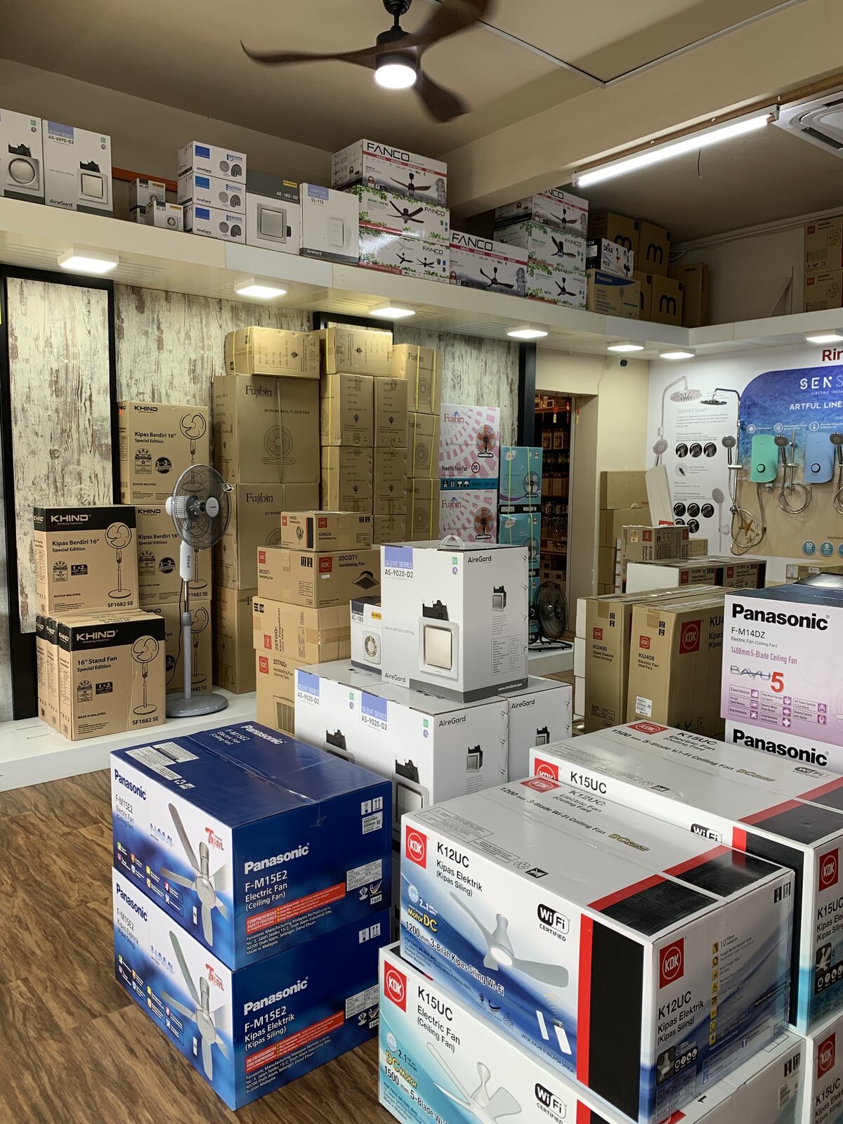 Stacks of boxed ceiling fans, ventilating fans and stand fans from several brands on a shop floor, with more fan boxes on a high shelf and a water-heater display wall behind