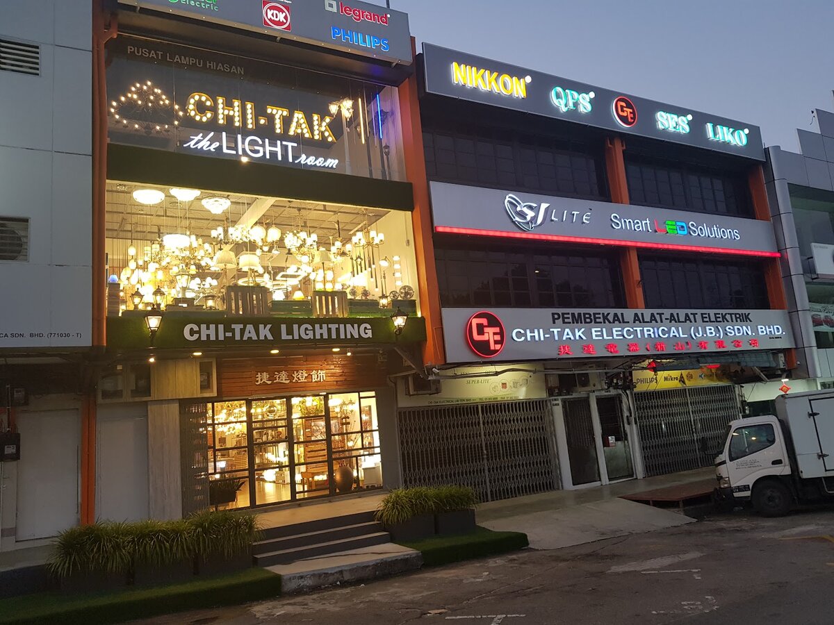 Two shophouses at dusk: a lit decorative-lighting showroom signed Chi-Tak Lighting on the left and a shuttered shop signed Chi-Tak Electrical (J.B.) Sdn Bhd with lighting brand logos on the right