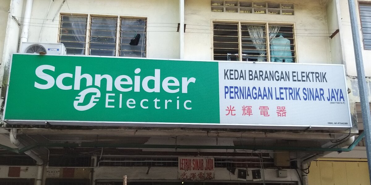 Shop fascia with a large green Schneider Electric panel and a white panel reading Kedai Barangan Elektrik, Perniagaan Letrik Sinar Jaya, with a small hand-painted sign over the doorway