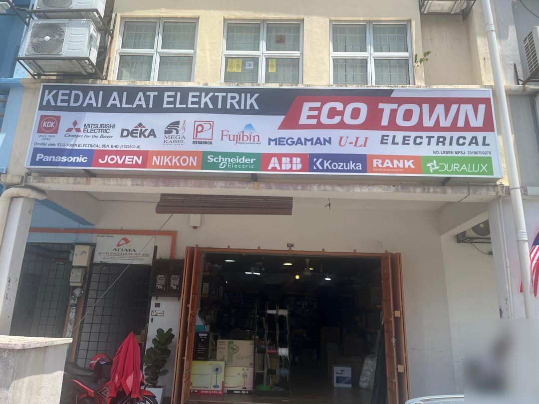 Shop unit with a long white signboard reading Kedai Alat Elektrik Eco Town Electrical and rows of brand logos, its folding doors open on a cable reel stand and boxed fans