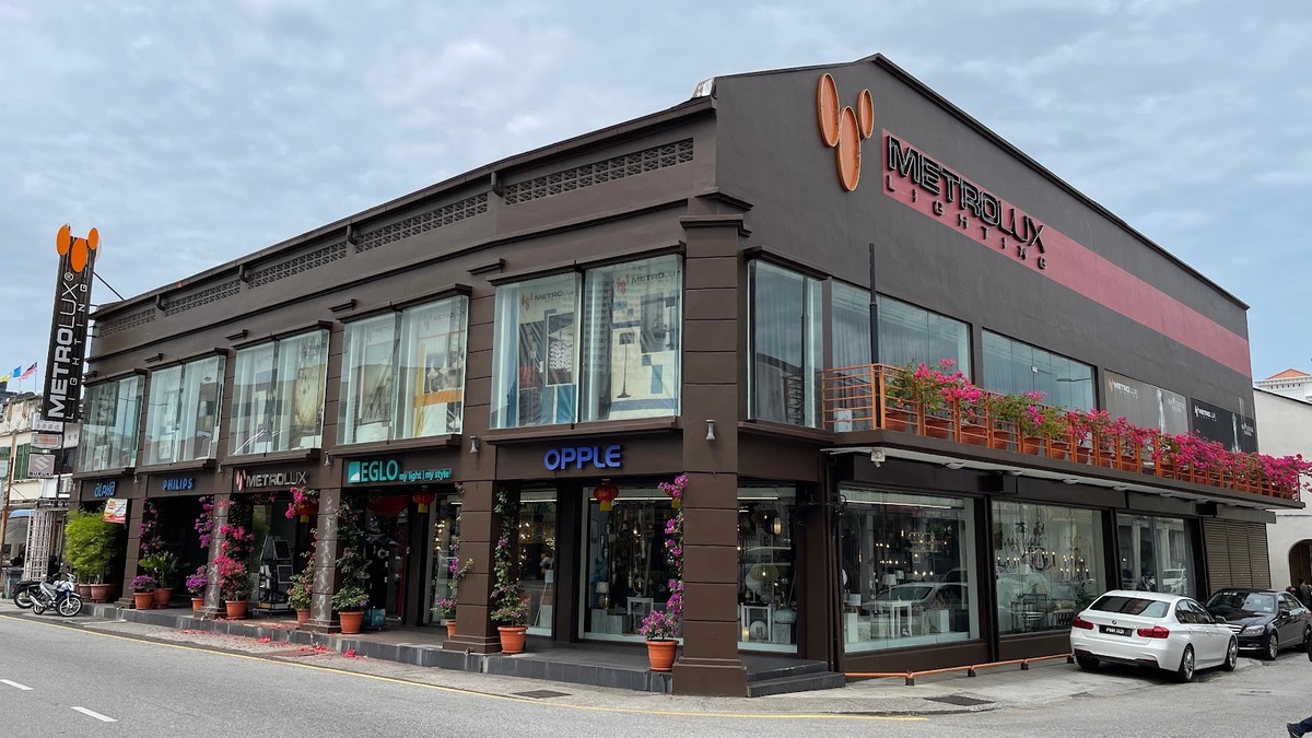 Two-storey brown corner shophouse row with brand signs above glass shopfronts and potted bougainvillea along the five-foot way