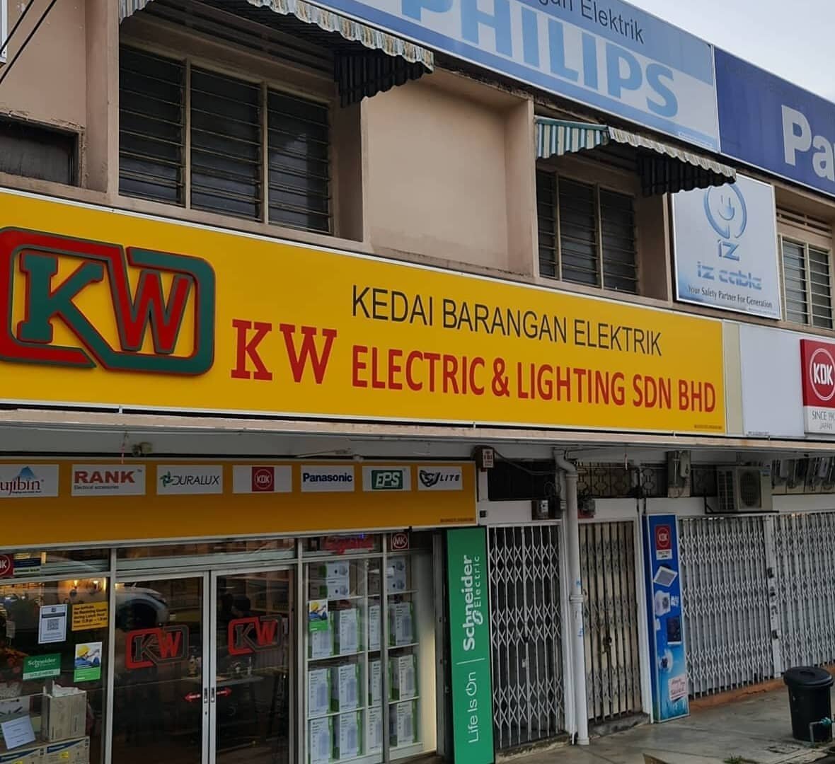 Corner shop with a yellow signboard reading Kedai Barangan Elektrik KW Electric & Lighting Sdn Bhd, brand logos below it, a Philips sign on the upper floor and a green Schneider Electric pillar sign beside glass doors
