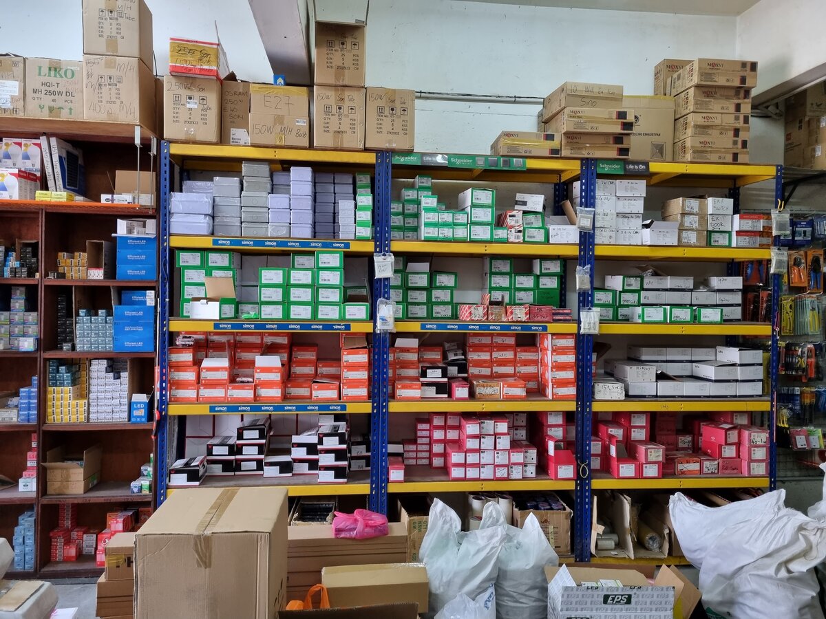 Steel shelving packed with green, white and orange boxes of switches and sockets, with brand strips on the shelf edges and stacked fan and lamp cartons above