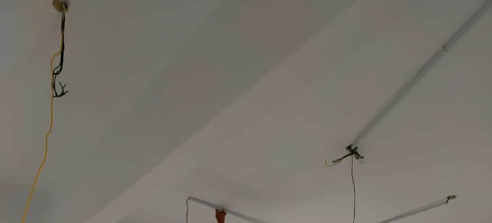 White ceiling with a cable drop on the left, a light point with loose wires and lengths of white PVC surface trunking running across it; a worker's hand at the bottom edge