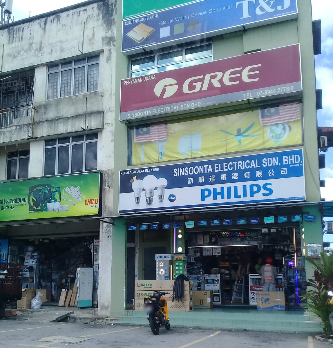 Shophouse with a blue and white signboard reading Sinsoonta Electrical Sdn Bhd and Philips, a red Gree sign and a blue T&J sign above, and an open shopfront with lamp and switch displays and K-Flex boxes