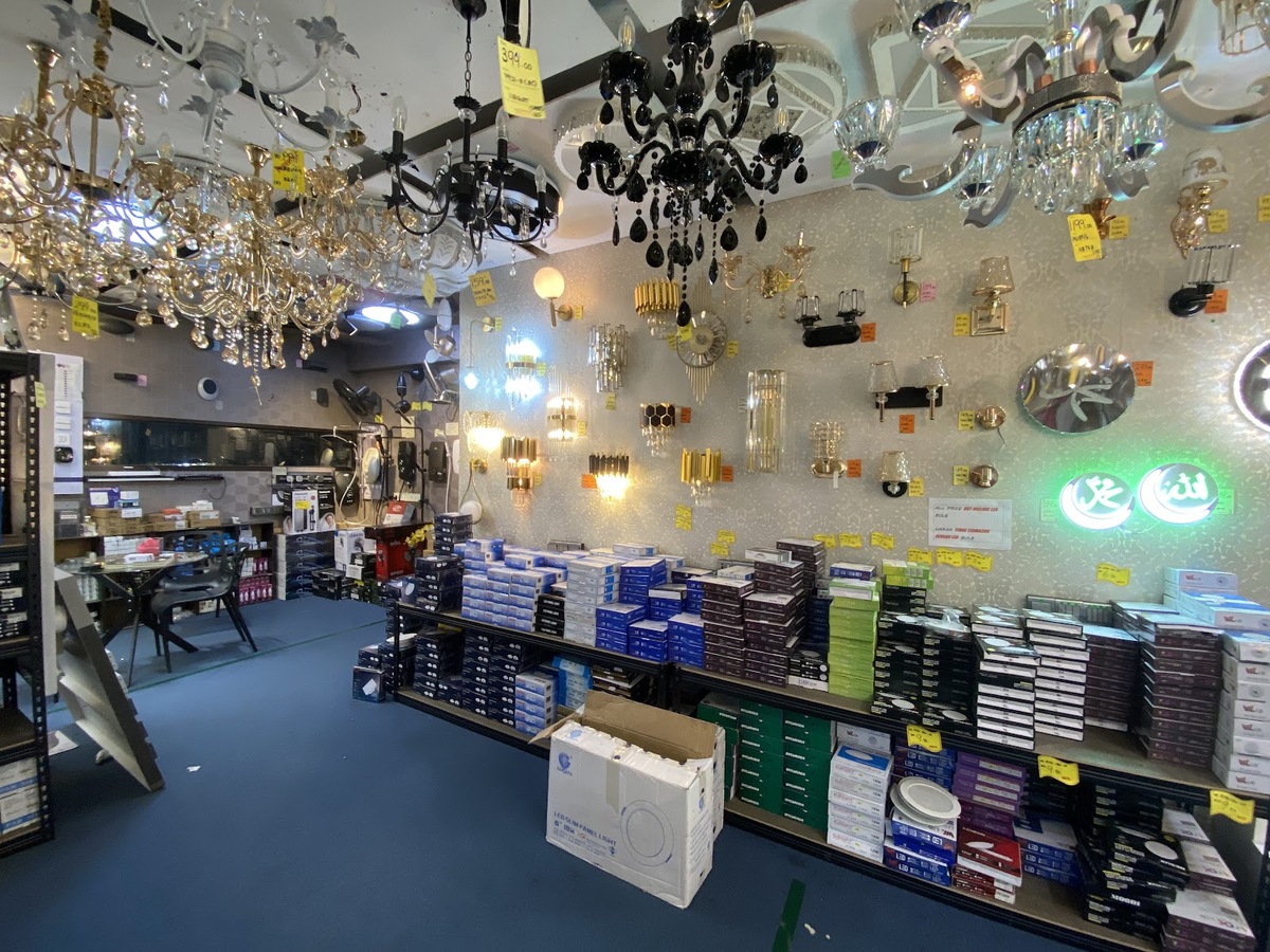 Shop interior with gold and black chandeliers hanging from the ceiling, a wall of wall lamps and long shelves stacked with lamp boxes