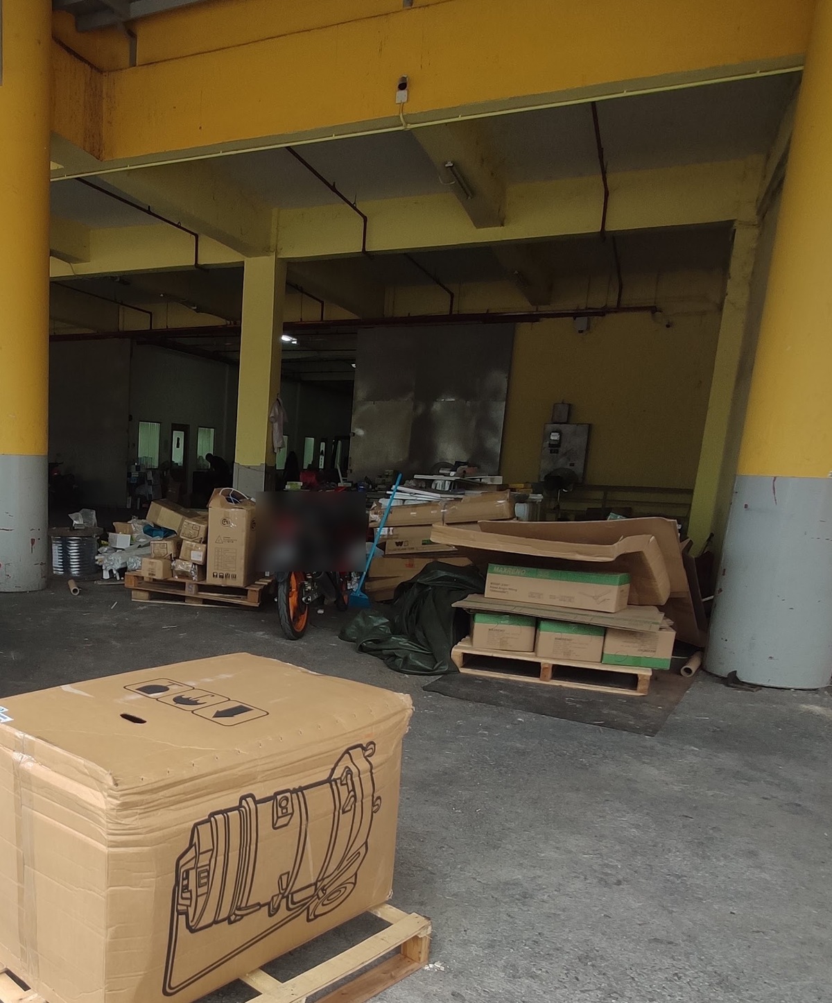 Large covered loading area with yellow concrete pillars, stacked cartons on wooden pallets and a big carton in the foreground