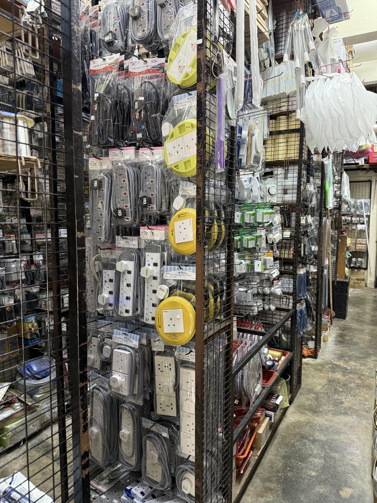 Wire-mesh racks hung with packaged grey and white extension cords and round yellow cable reels, with adaptors, timer switches and boxes of plug tops on racks behind