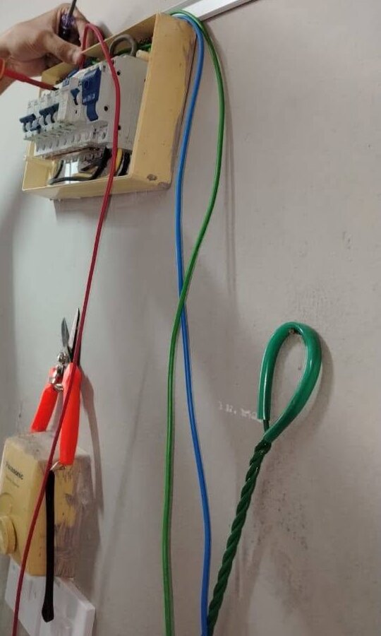 Small cream-coloured distribution board with a row of breakers on a wall, hands holding a screwdriver at the top, red, blue and green leads hanging down past a socket box and a looped green cable