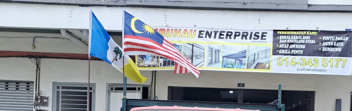 Ground floor of a shop row with Penang and Malaysian flags in front of a long banner reading Embunau Enterprise, a list of metalwork services and the number 014-343 5177, above an open shop entrance numbered 32