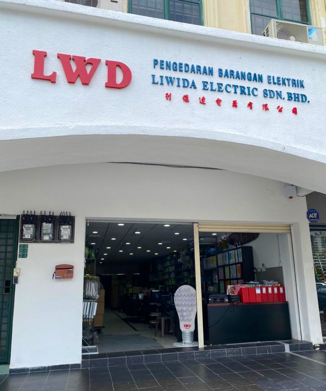 White shophouse front with a large red LWD logo and blue lettering reading Pengedaran Barangan Elektrik Liwida Electric Sdn Bhd, above an open shop entrance with a bulb-shaped LWD display