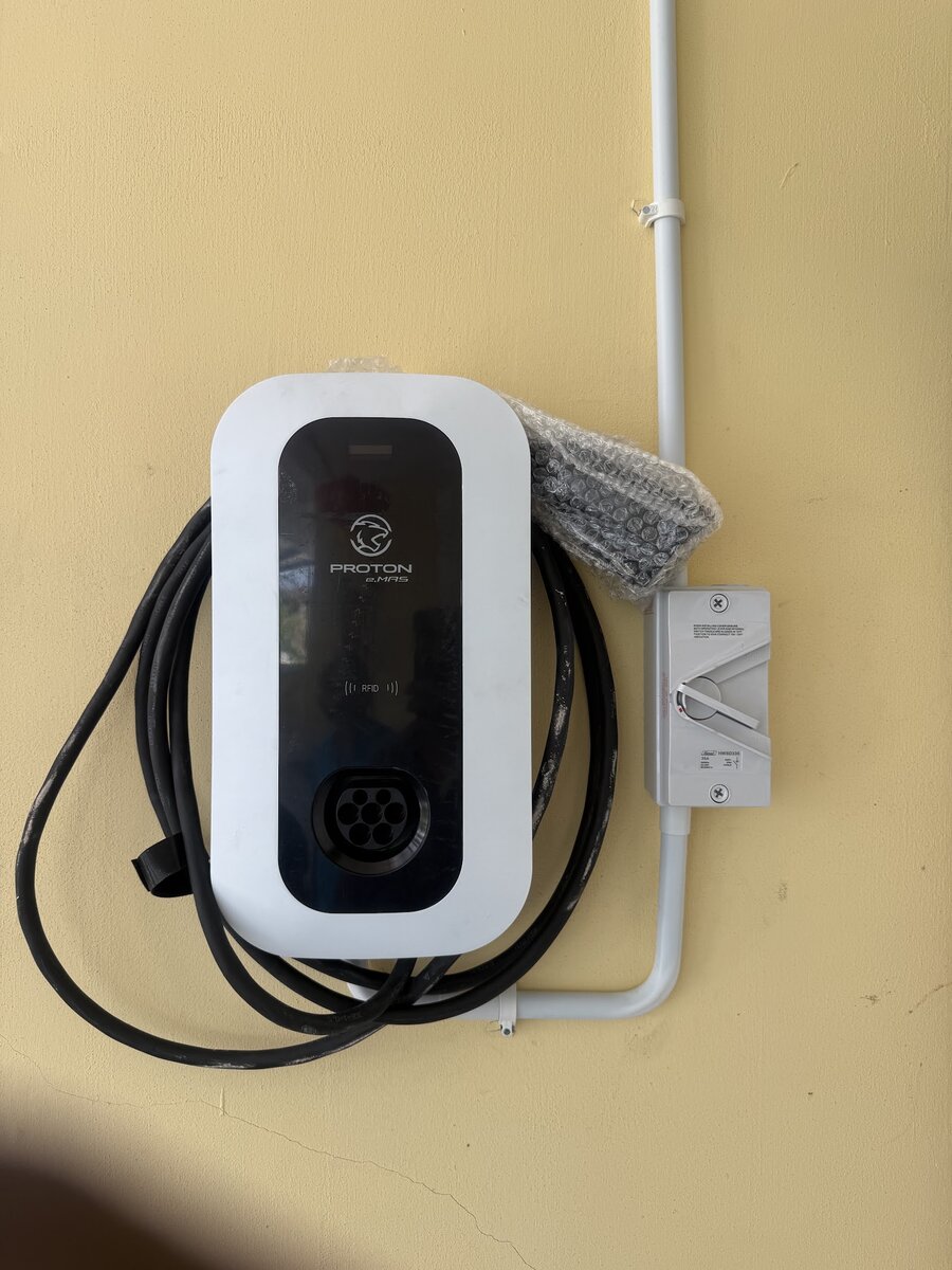 White Proton e.MAS EV wallbox with a coiled charging cable, mounted on a yellow wall next to a grey isolator switch fed by conduit