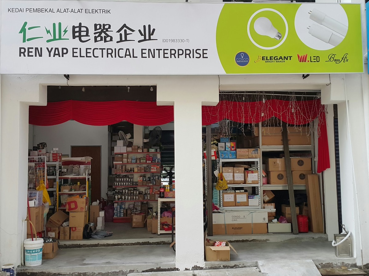 Shopfront with a green and white signboard reading Ren Yap Electrical Enterprise, Kedai Pembekal Alat-Alat Elektrik, above an open shop with shelves of boxed lights