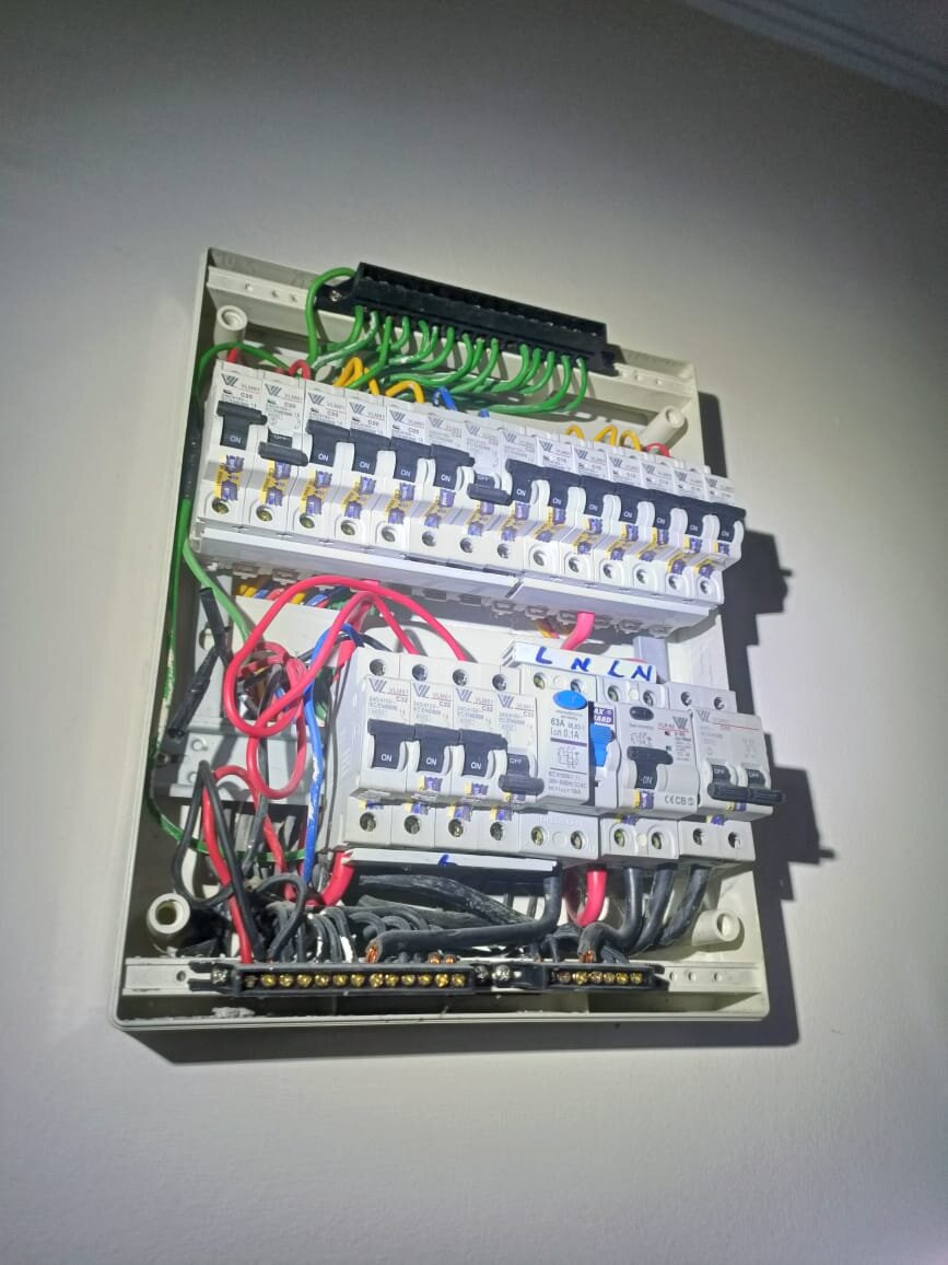 An open cream distribution board on a white wall: a black terminal block with green earth wires at the top, a row of white MCBs, and a lower row with more MCBs, two RCCBs (one with a blue test button) and a main switch under a hand-written L N L N label