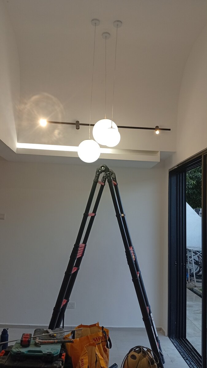Tall white room with a cove-lit ceiling, three glass globe pendant lights and a black bar light hanging from it, and an A-frame ladder below