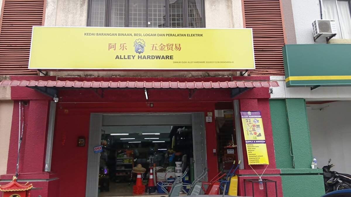 Red shopfront with a yellow "Alley Hardware 阿乐五金贸易" signboard, traffic cones and wheelbarrows at the entrance