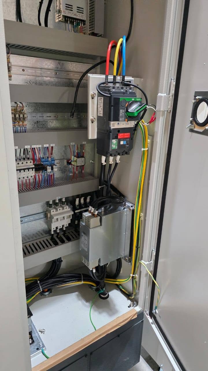 Tall grey control cabinet with its door open: a black moulded-case circuit breaker fed by red, yellow and blue cables, rows of terminal blocks and small breakers, and a grey filter unit
