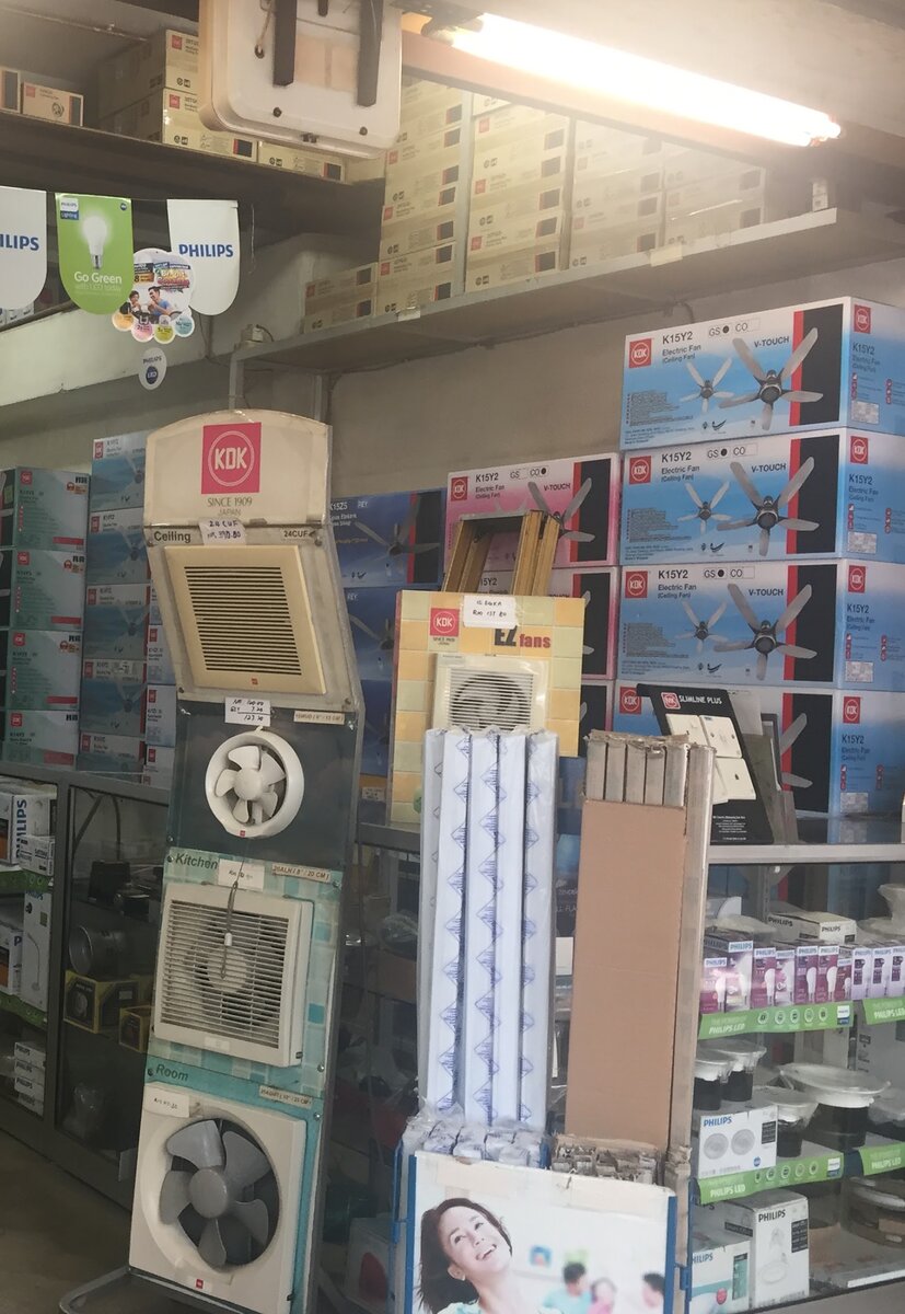 Shop interior with stacks of blue and pink KDK fan boxes, a stand of wall-mounted ventilating fans, Philips pennants overhead and a glass counter of boxed lamps