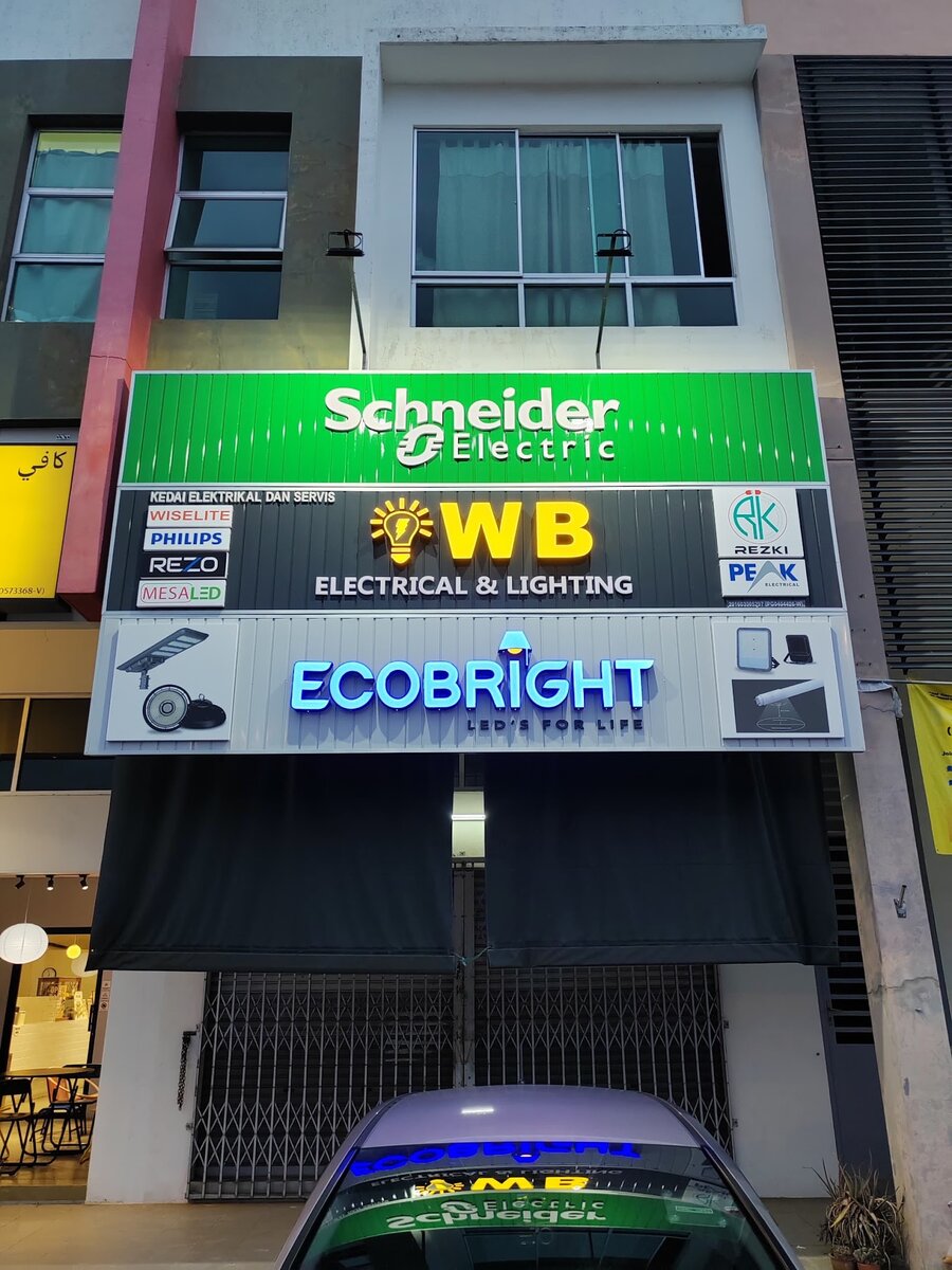 Shophouse front at dusk with a green Schneider Electric lightbox, a dark WB Electrical & Lighting signboard and a blue Ecobright sign above closed shutters