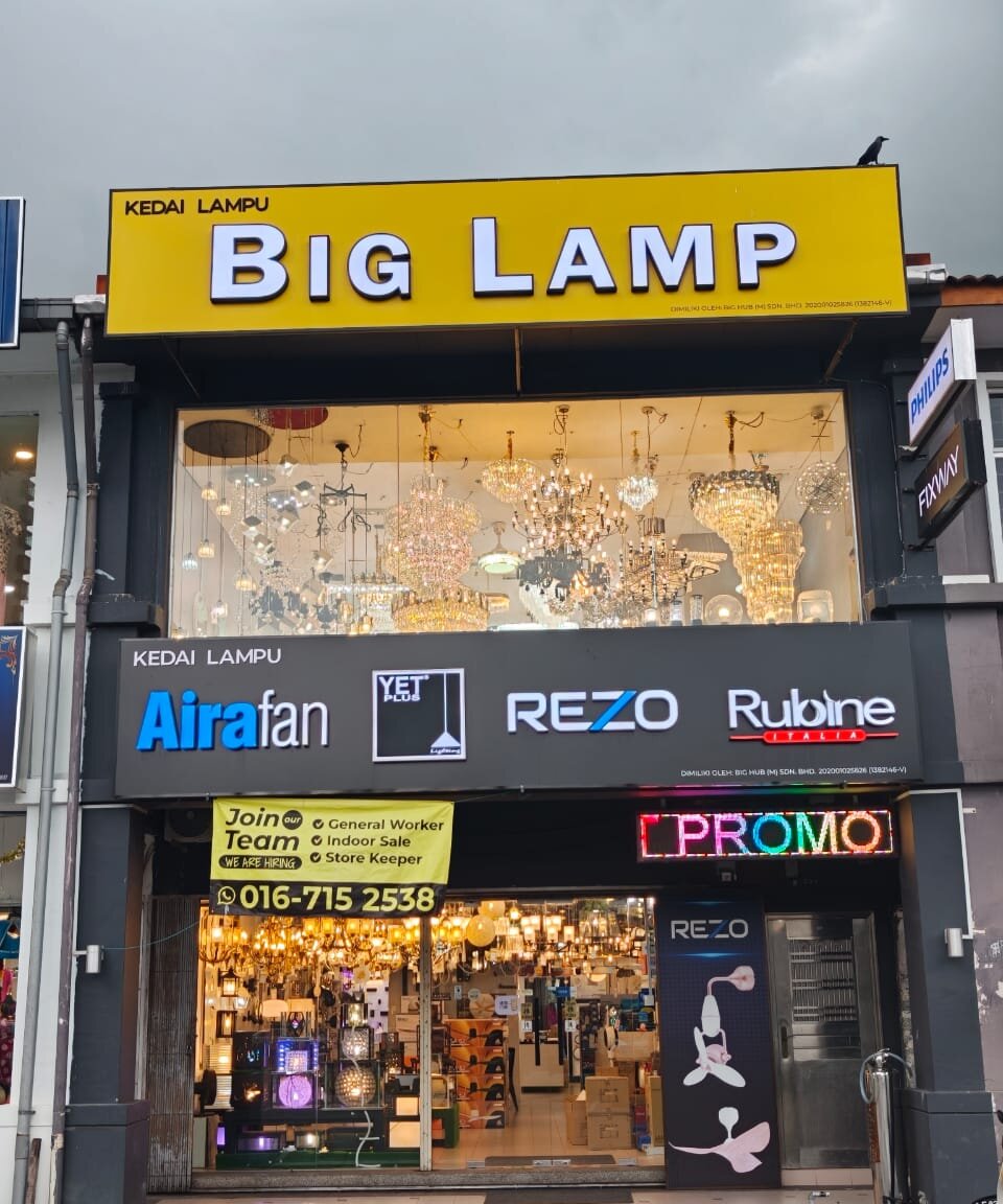 Two-storey shopfront with a yellow Big Lamp sign, a dark signboard with fan and lighting brand logos and a lit showroom of chandeliers